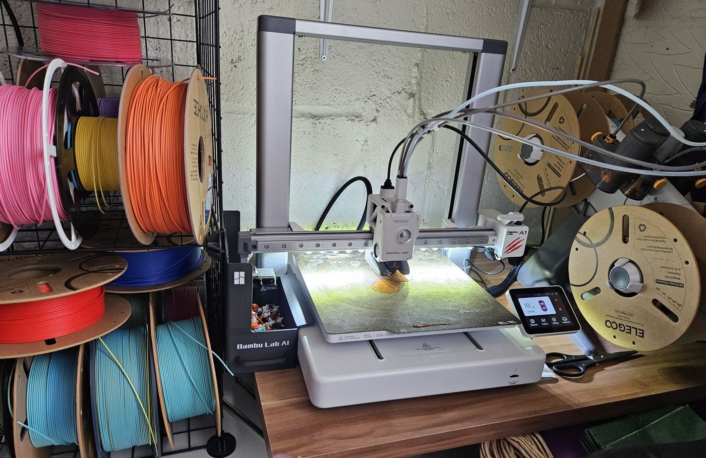 The Hark Tech 3D printing bench — Bambu Lab A1 with AMS, full filament rack on the left in pink, orange, yellow, red and teal, K2 out of frame to the right